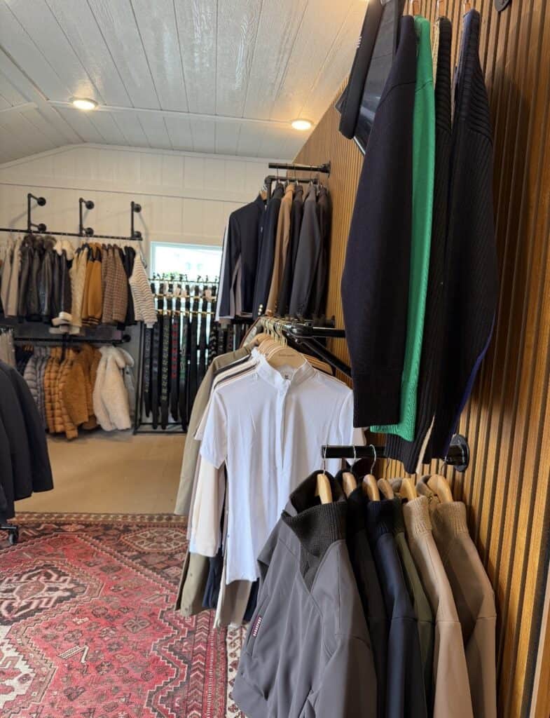 Clothing racks filled with jackets, sweaters, and shirts in a retail store with a patterned rug on the floor.