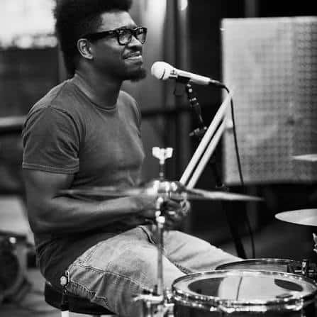 A man wearing glasses plays the drums and sings into a microphone in a black and white indoor setting.