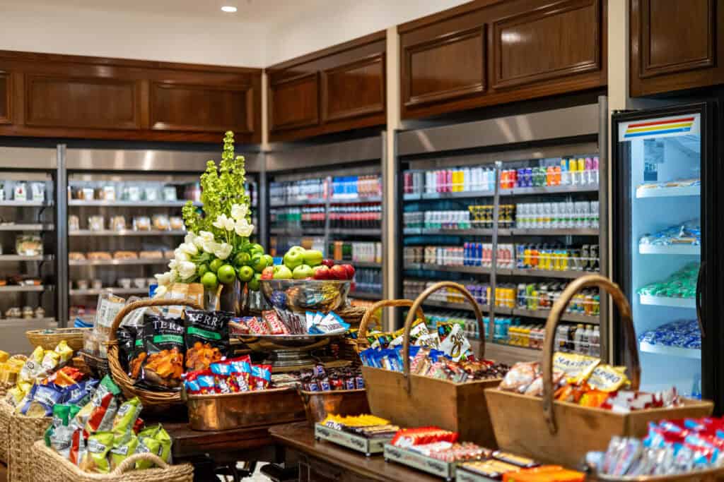 A well-lit convenience store interior with shelves and baskets filled with snacks, and refrigerators stocked with drinks in the background.