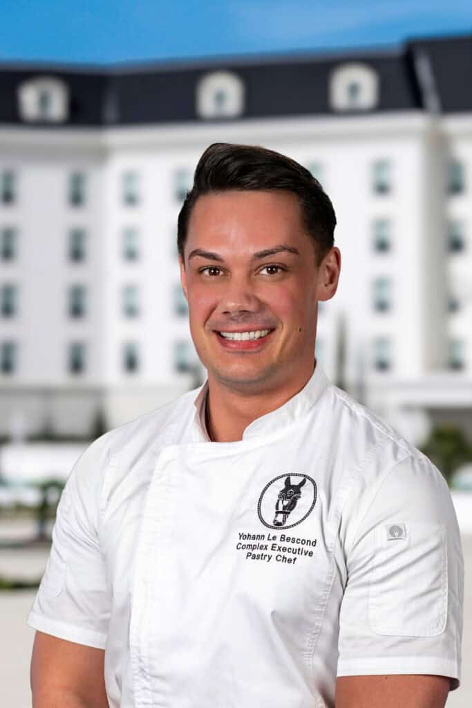 A chef in a white uniform stands outdoors in front of a large white building, smiling at the camera.