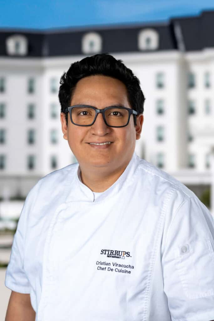 A man wearing glasses and a white chef's coat stands outdoors in front of a large white building with many windows.