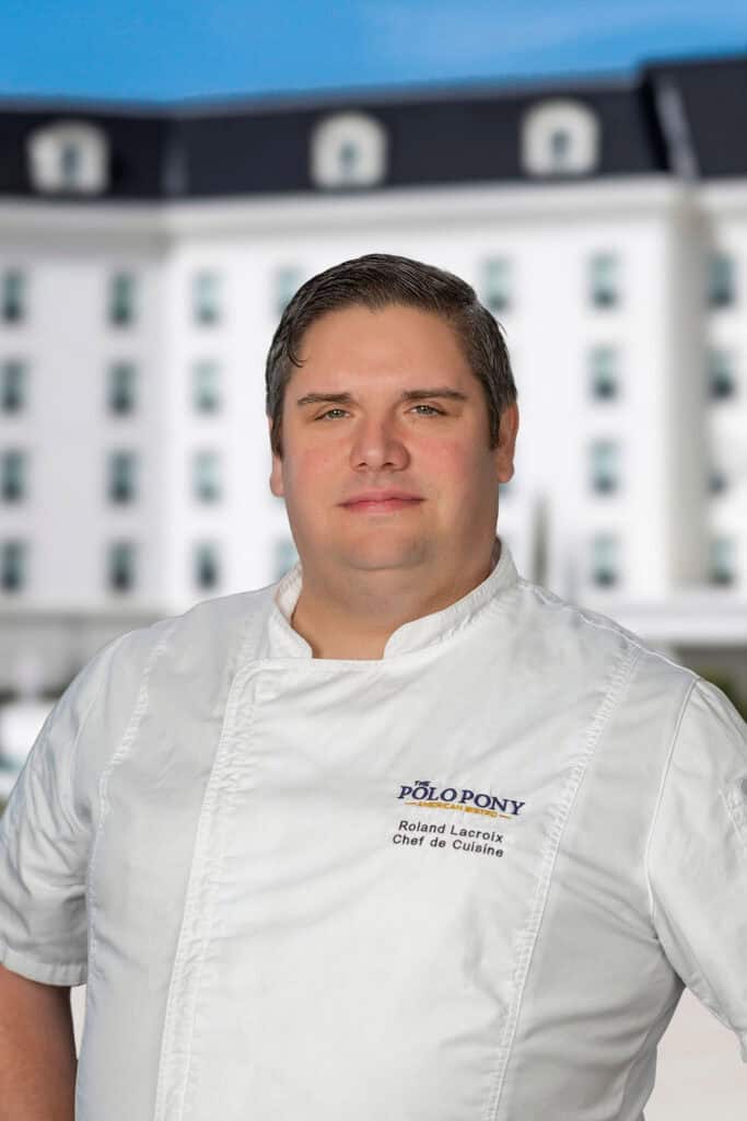 A chef in a white uniform stands outdoors with a blurred white building in the background. The chef’s jacket has “Polo Pony” and other text embroidered on it.