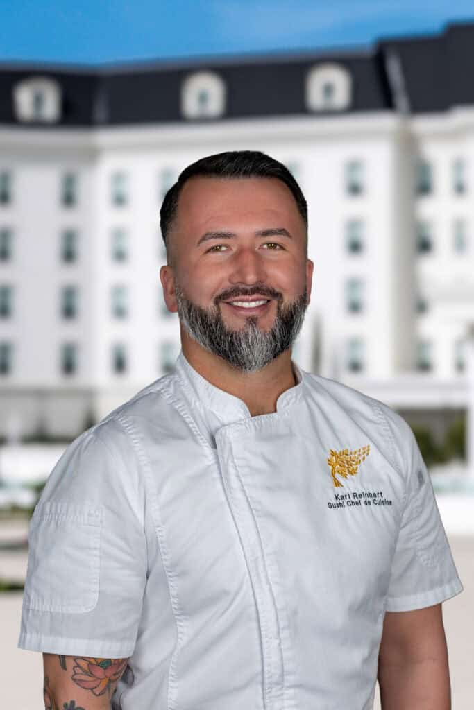 A man with a beard in a white chef’s coat stands outside a large white building, smiling at the camera.
