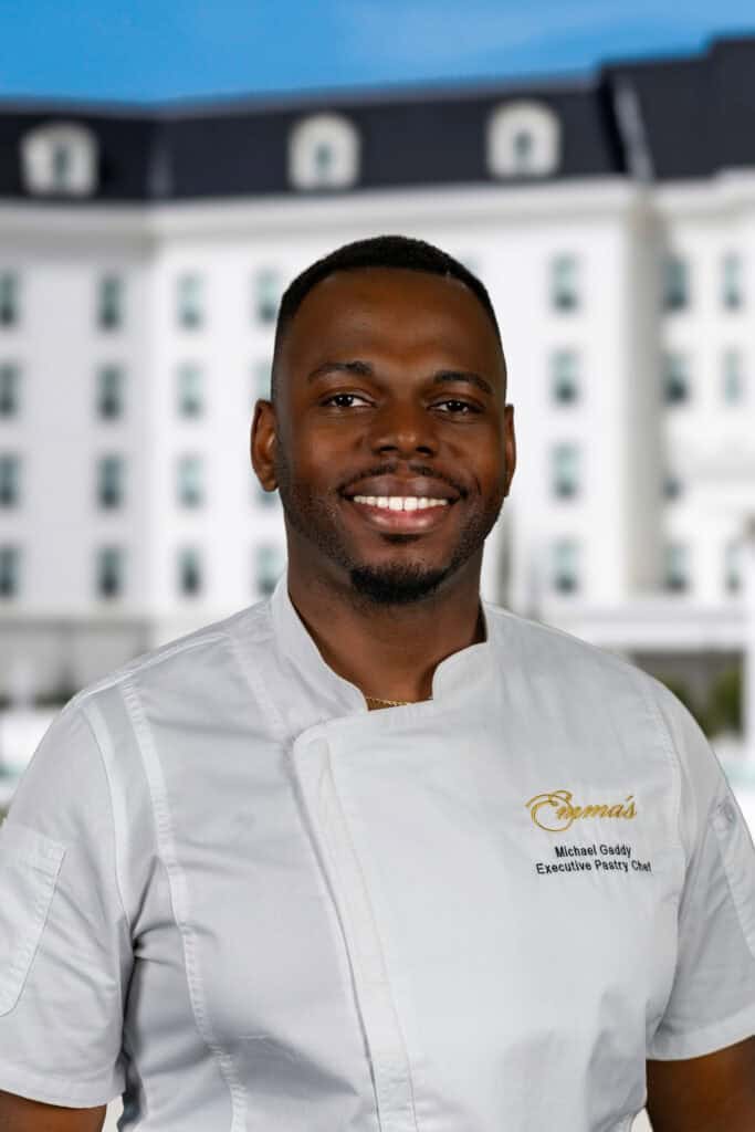 A man in a white chef's jacket stands smiling in front of a blurred white building. The jacket reads: “Emma’s, Michael Gaddy, Executive Pastry Chef.”.