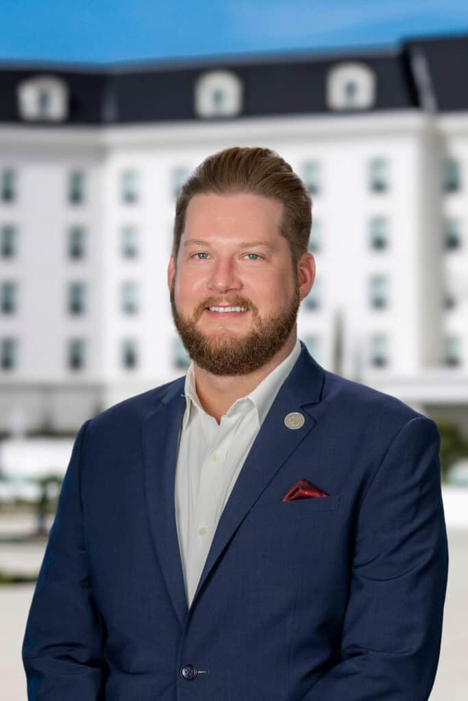 A man in a blue suit with a white shirt and red pocket square stands outside in front of a large white building with dark roof accents.