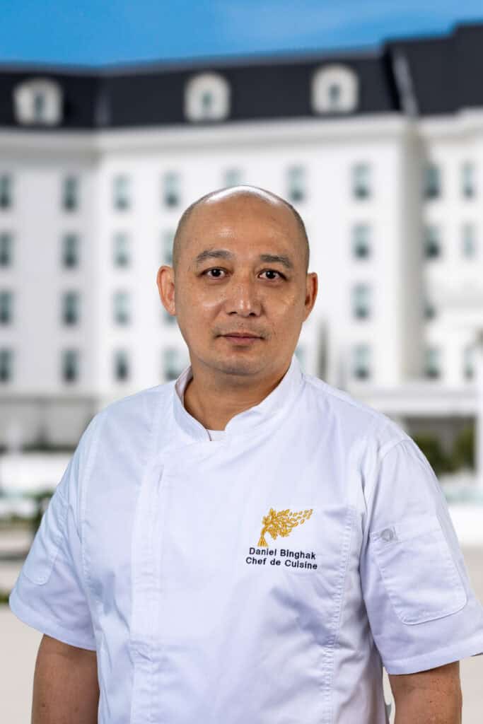 A chef in a white uniform stands outdoors in front of a large, white building.