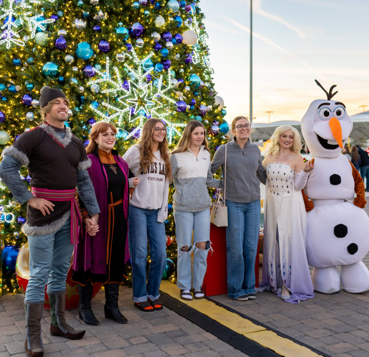 Six people, including two in “Frozen” character costumes and one in an Olaf mascot suit, stand in front of a large decorated Christmas tree outdoors at sunset.
