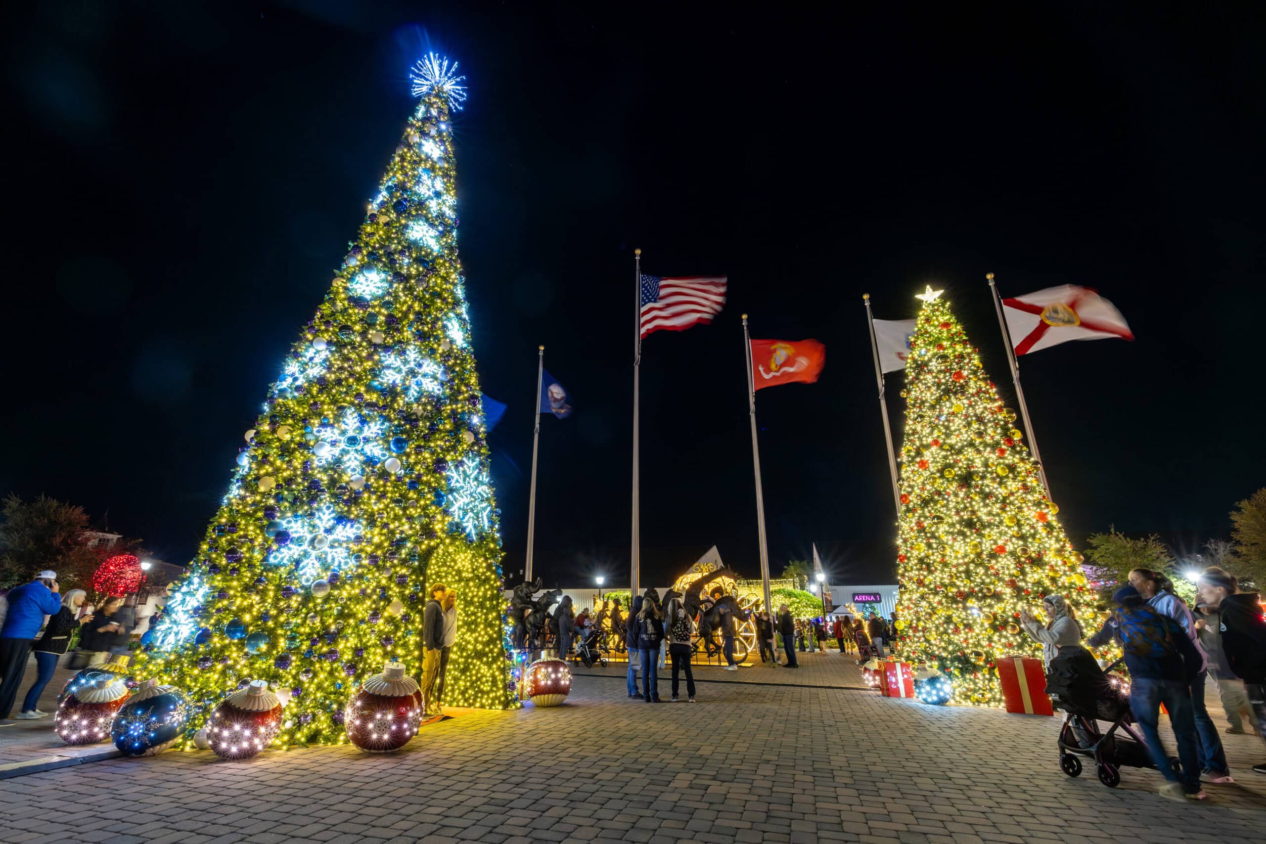 Two large decorated Christmas trees with lights stand in a plaza at night, with people gathered around and flags flying in the background.
