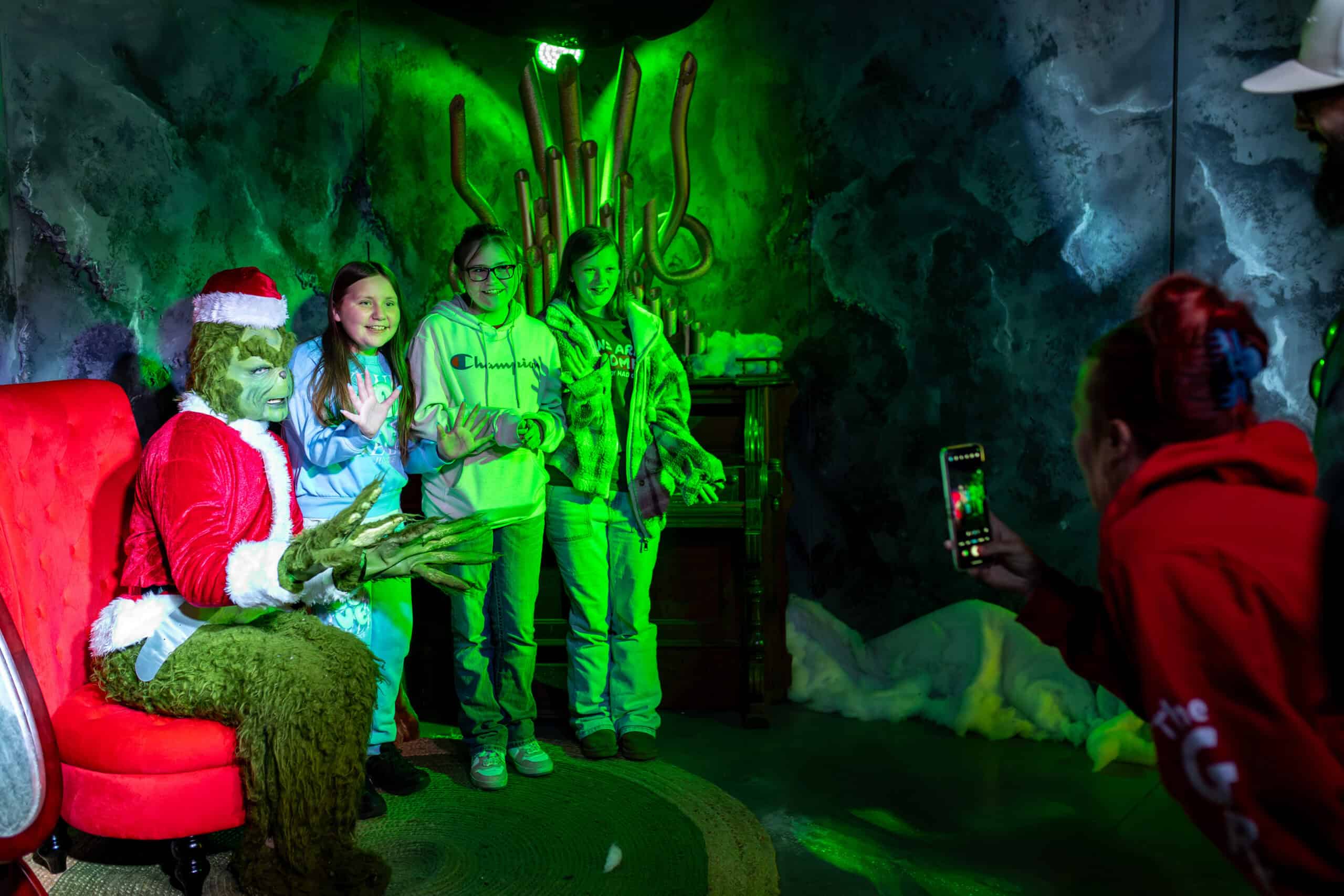 Three kids pose for a photo with a person in a Grinch costume dressed as Santa, while another person takes their picture in a room lit with green light.