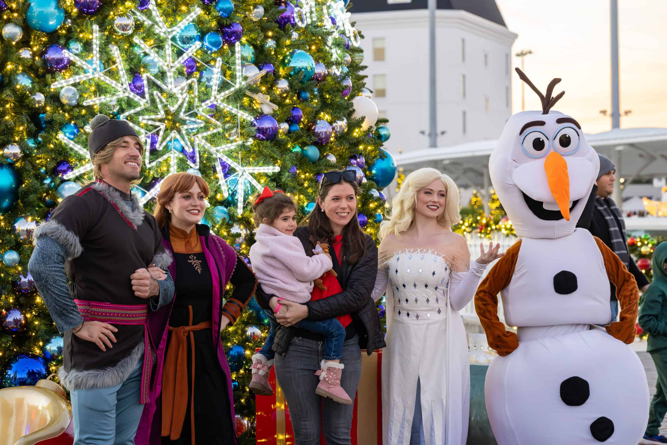 A group poses in front of a decorated Christmas tree, including people in costumes resembling characters from "Frozen" and a woman holding a child.