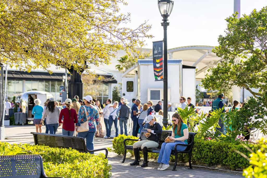 People gather at an outdoor market on a sunny day; some sit on benches while others stand in line near vendor trucks in a park-like setting.