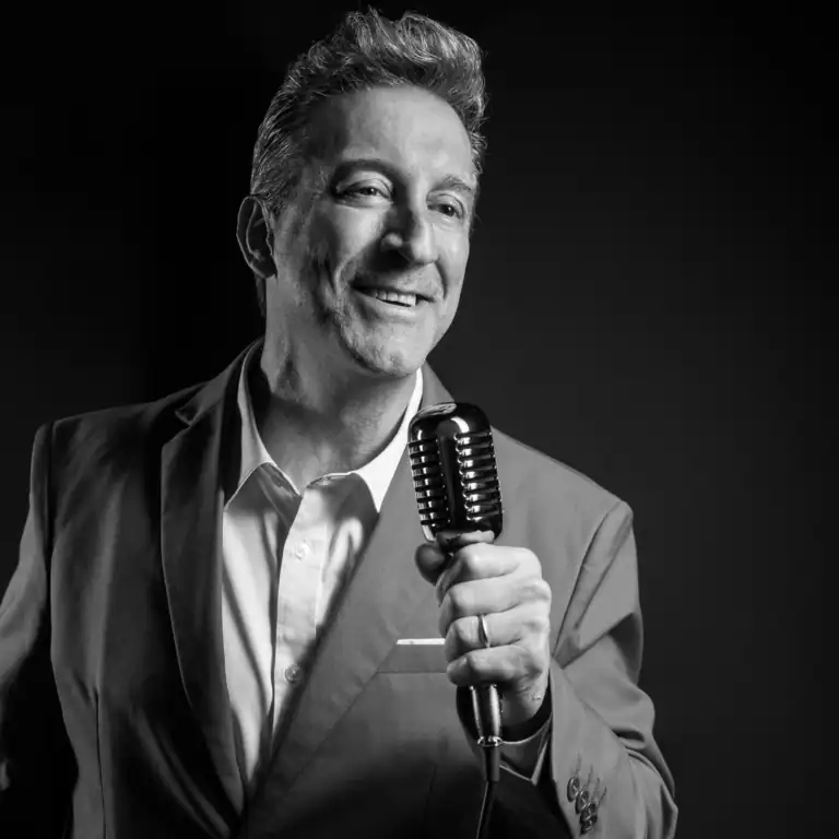 A man in a suit holds a vintage microphone and smiles while looking slightly to the side, standing against a dark background. The image is in black and white.