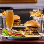 A large cheeseburger with lettuce, tomato, onion, and pickles is served on a plate with fries and a glass of beer on a restaurant table.