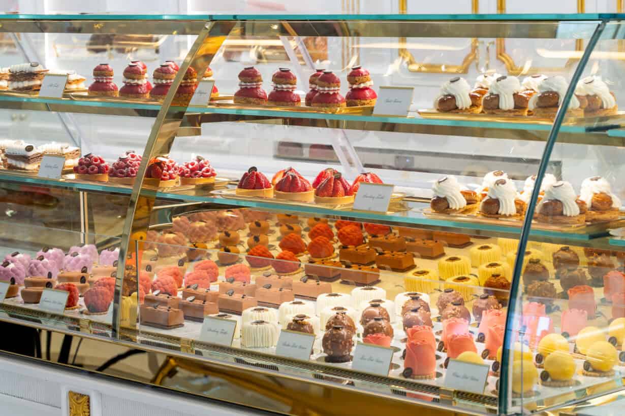 A glass display case filled with assorted colorful pastries, tarts, cakes, and desserts arranged on multiple shelves in a bakery or patisserie.