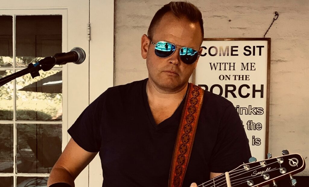 A man wearing sunglasses and a black t-shirt plays an acoustic guitar in front of a microphone, with a sign in the background inviting people to sit on the porch.