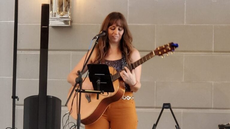 A woman plays an acoustic guitar and sings into a microphone, standing in front of a beige tiled wall.