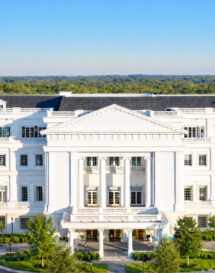 Large white neoclassical building with columns and multiple windows, surrounded by trees under a clear blue sky.