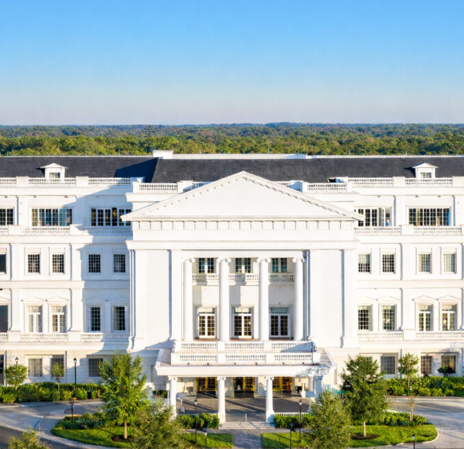 Large white neoclassical building with columns and multiple windows, surrounded by trees under a clear blue sky.