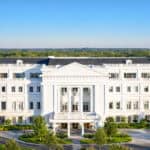 A large white neoclassical building with columns and multiple windows, surrounded by trees under a clear blue sky.