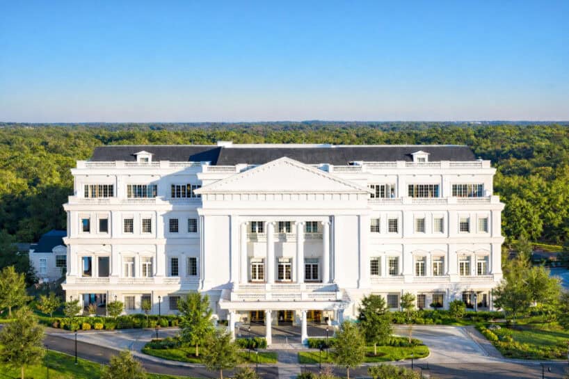 A large white neoclassical building with columns and multiple windows, surrounded by trees under a clear blue sky.