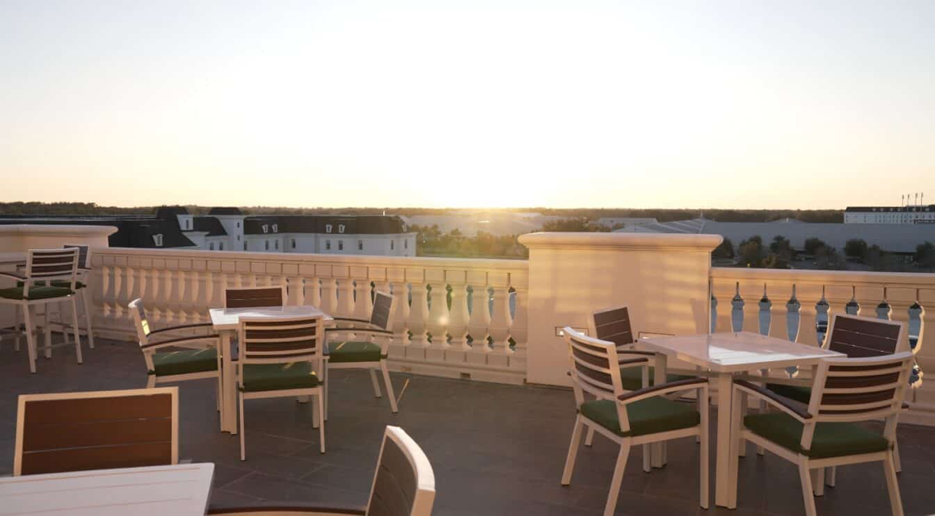 Outdoor rooftop terrace with white tables and chairs, green cushions, and a white balustrade, overlooking buildings at sunset.