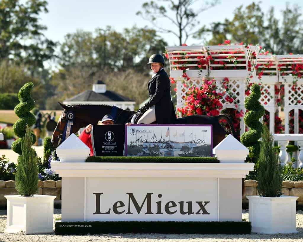 A smiling equestrian sits on a horse behind a large ceremonial check and a podium labeled "LeMieux" at an outdoor event with flowers and topiary decorations.