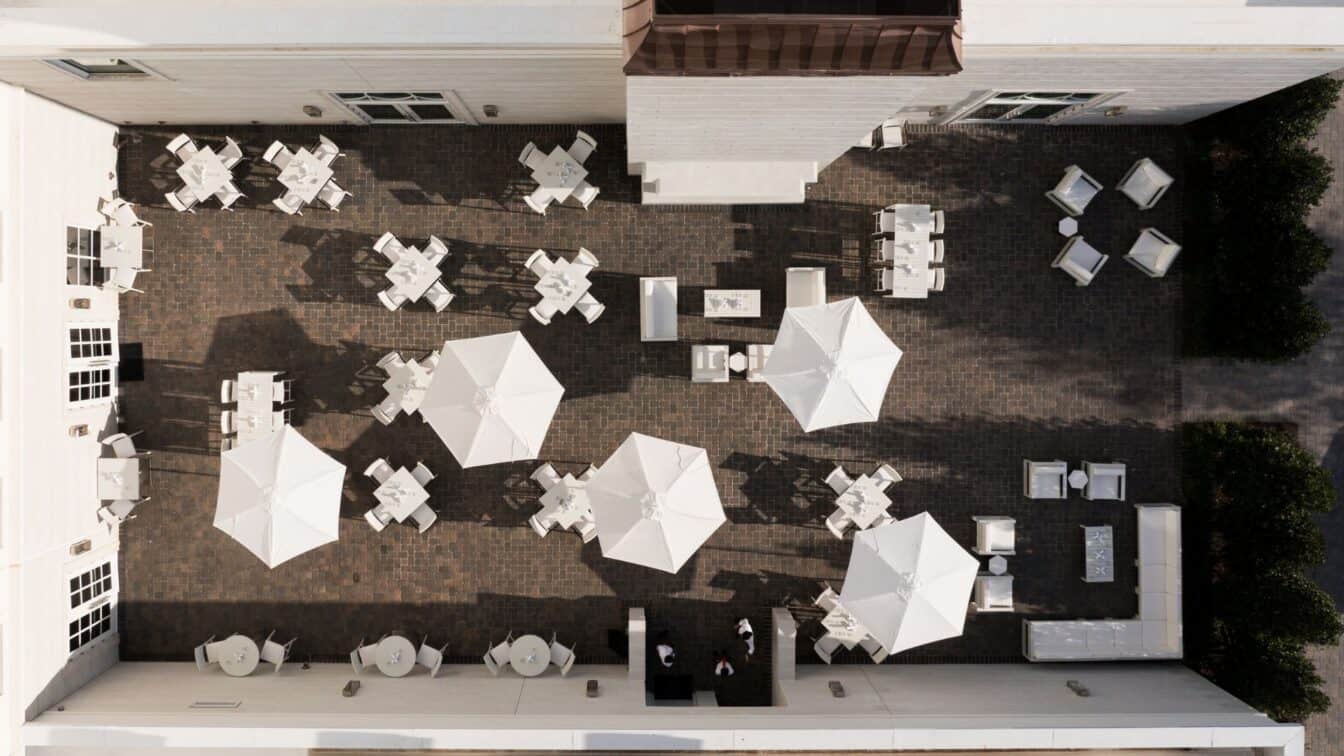 Aerial view of an outdoor patio with tables, chairs, and umbrellas arranged on a paved surface next to a white building.