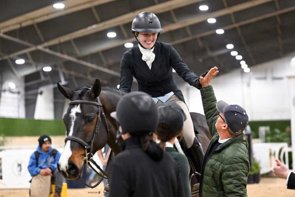 A smiling equestrian on horseback gives a high-five to a standing man inside an indoor riding arena, with other people present in the background.