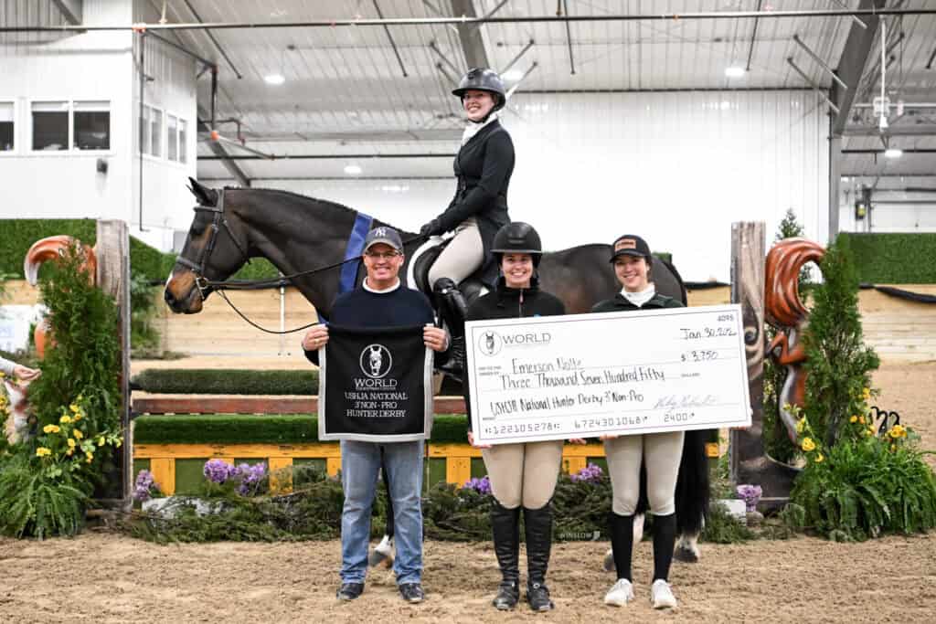 Four people pose with a horse in an indoor arena; one person sits on the horse while the others hold a large check and a winner's plaque.