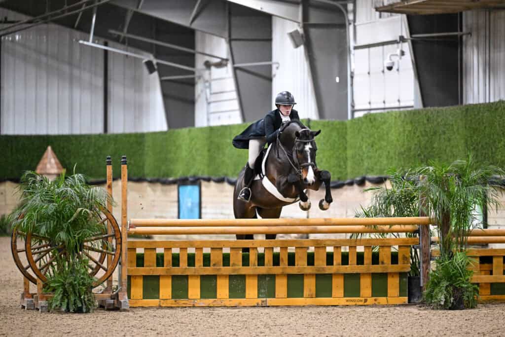 An equestrian rider in formal attire jumps a horse over a wooden obstacle in an indoor arena with greenery and decorative wagon wheels.