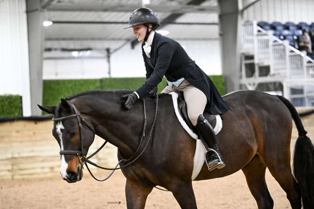 A rider wearing a helmet and formal riding attire pats a dark horse while riding in an indoor equestrian arena.