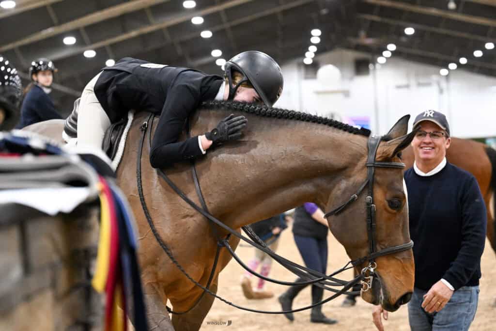 A rider hugs a horse, leaning forward in the saddle, while a man in a hat and glasses stands nearby, smiling, in an indoor equestrian arena.