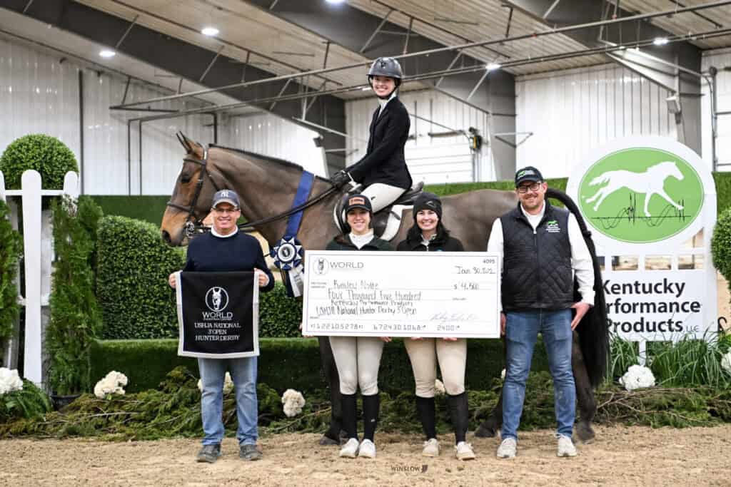Five people pose indoors with a horse, a large presentation check, and an award banner in an equestrian competition setting with green and white decor.