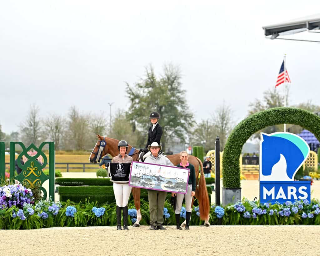 Three people and a horse stand in an arena holding a large award check, with floral decorations, event signage, and a US flag in the background.