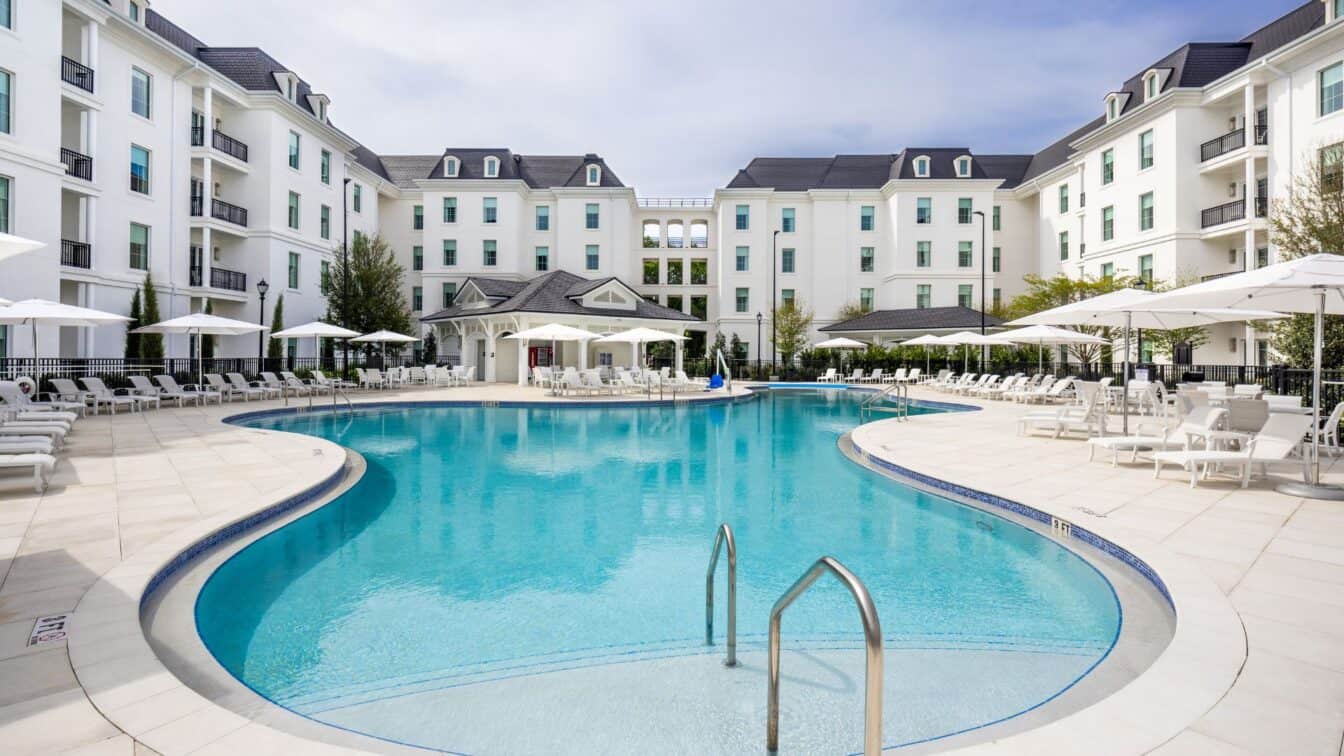 Curved outdoor swimming pool surrounded by white lounge chairs, umbrellas, and a multi-story white residential building in the background.