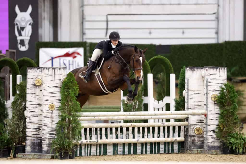 A person on horseback jumps over a white gate obstacle during an indoor equestrian event.
