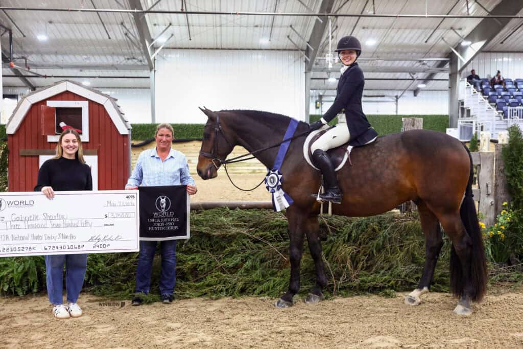 Three people at an indoor horse arena: one holding a large check, one holding a plaque, and one riding a horse wearing a ribbon.