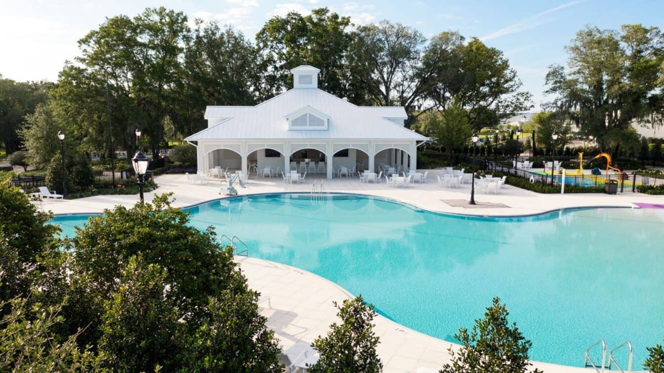 Large outdoor swimming pool with clear blue water, surrounded by trees and lounge chairs, in front of a white pavilion under a partly cloudy sky.