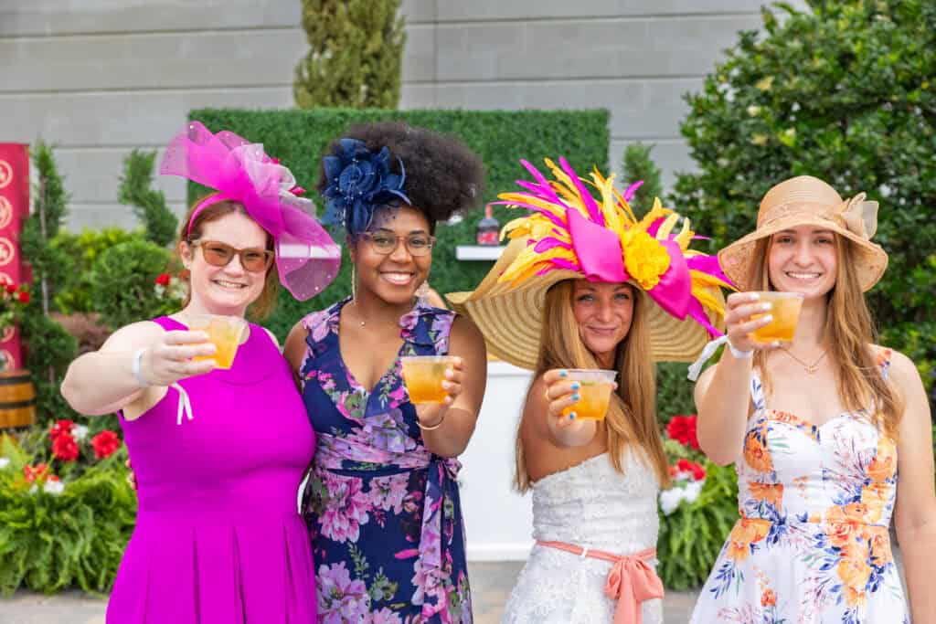 Four women in colorful dresses and decorative hats stand outdoors, smiling and holding up drinks, with greenery and flowers in the background.