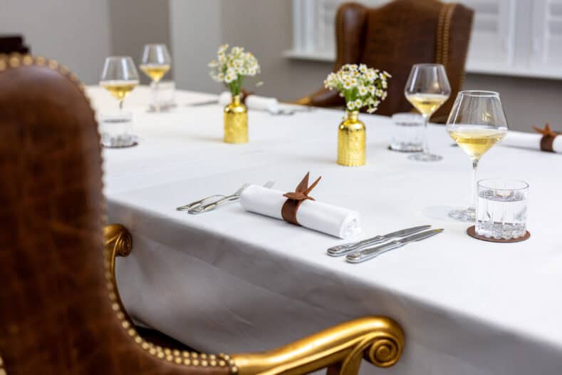 Elegant table setting with white tablecloth, rolled napkins with leather rings, wine glasses, water glasses, and small gold vases with flowers on a formal dining table.