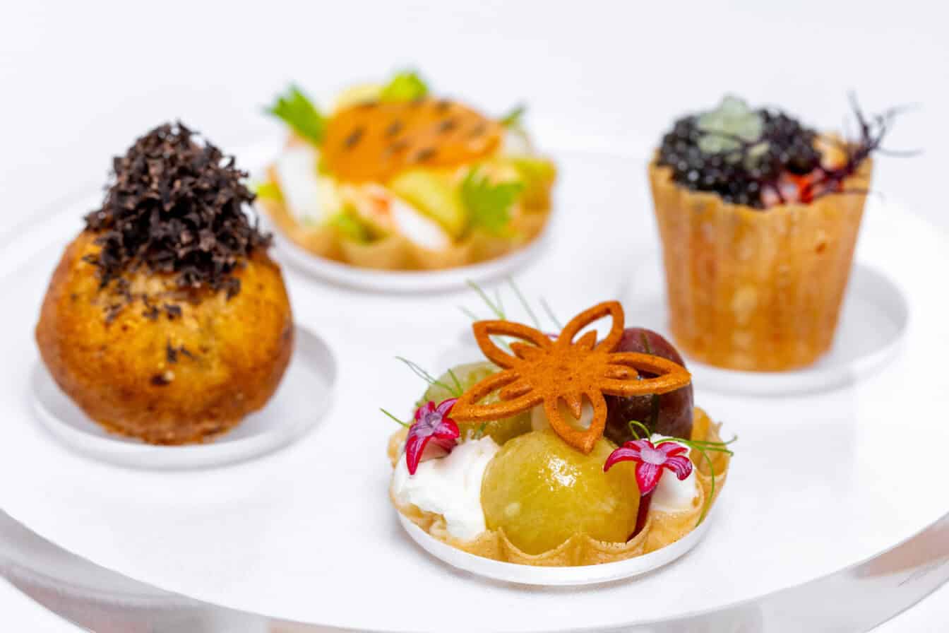 A selection of four assorted gourmet bite-sized appetizers, including tarts and a pastry, each garnished with herbs, edible flowers, and toppings, displayed on a white plate.