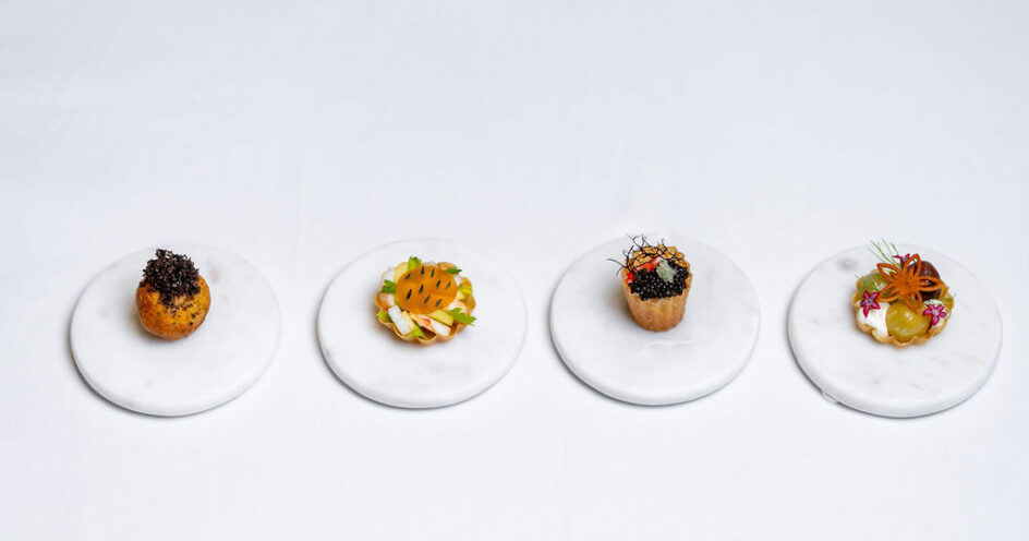 Four assorted gourmet appetizers are displayed on separate round white marble plates, spaced evenly in a row on a plain white surface.