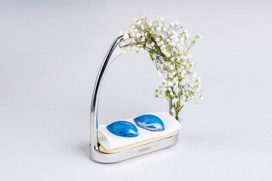 A silver stand holds two blue stones on a white cushion, adorned with small white flowers on the upper right side.