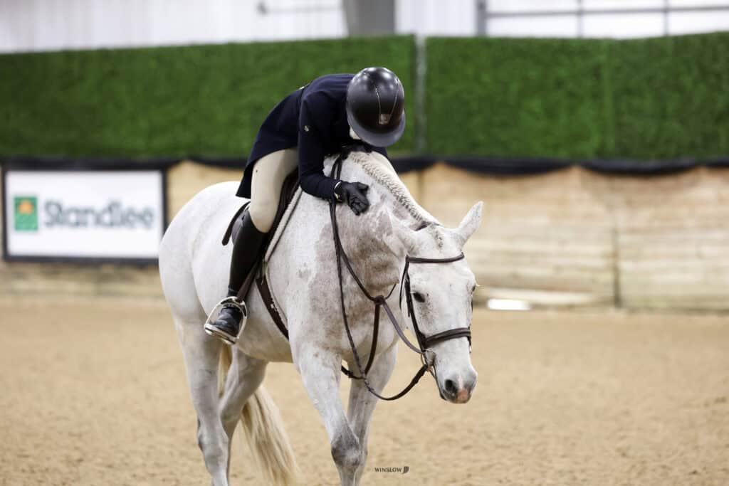 A rider in equestrian attire leans forward to hug a white horse in an indoor arena.