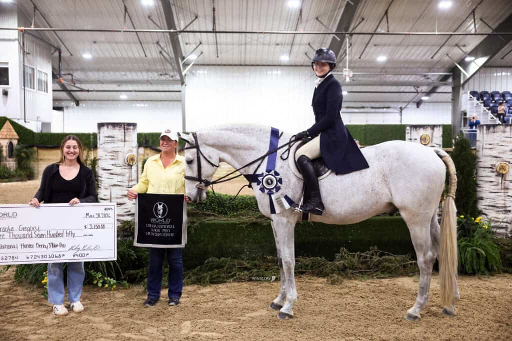 A woman on horseback poses with two others, one holding a large check and the other a gift bag, in an indoor equestrian arena with decorative fences in the background.