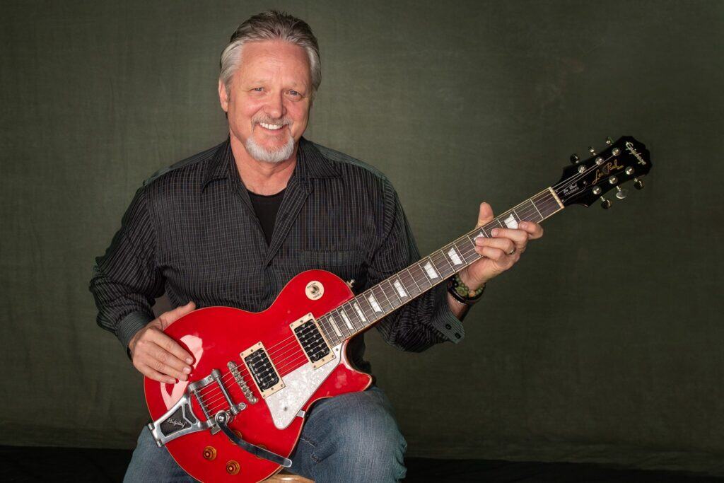 A man with gray hair and a beard sits holding a red electric guitar, smiling at the camera against a plain dark background.