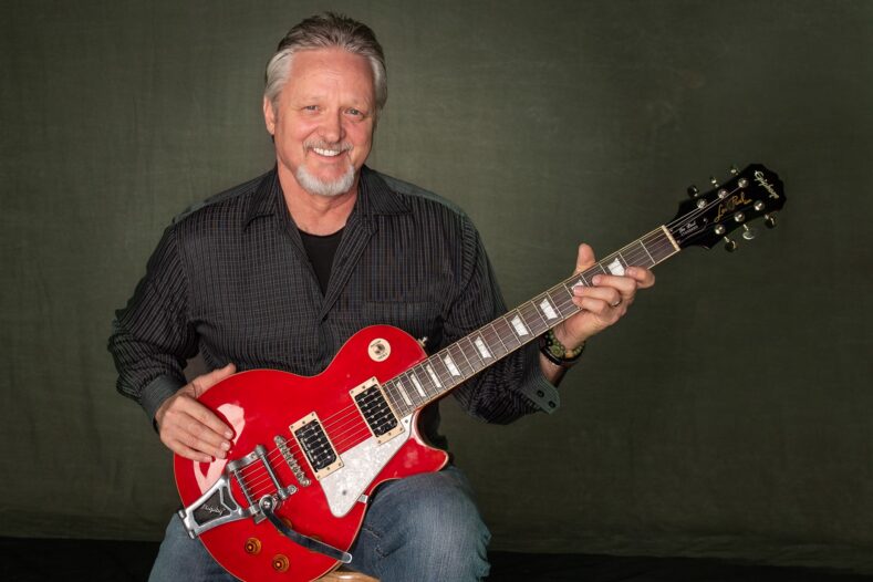 A man with gray hair and a beard sits holding a red electric guitar, smiling at the camera against a plain dark background.