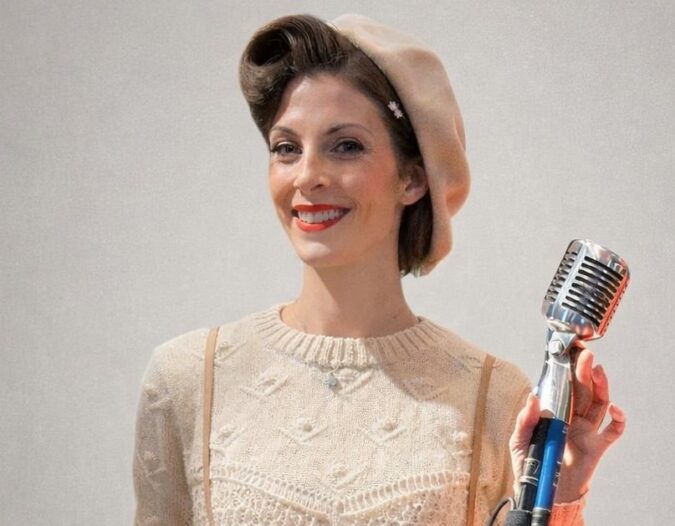 Woman in vintage-style clothing and beret stands smiling while holding a retro microphone against a plain background.
