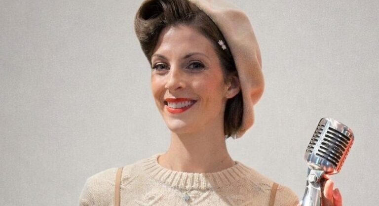 Woman in vintage-style clothing and beret stands smiling while holding a retro microphone against a plain background.