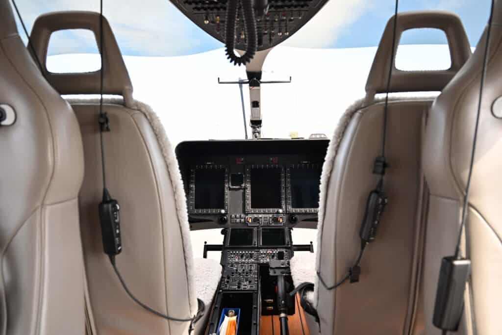 View of a modern helicopter cockpit from behind the pilot and co-pilot seats, showing flight controls, instrument panels, and headsets.